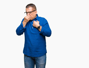 Middle age arab man wearing glasses over isolated background smelling something stinky and disgusting, intolerable smell, holding breath with fingers on nose. Bad smells concept.
