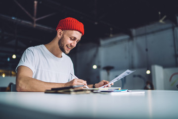 Handsome Caucasian man in trendy red cap making memo notes during learning process for preparing to...