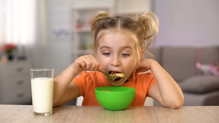 Happy little girl eating cornflakes with milk sitting home table, healthy food