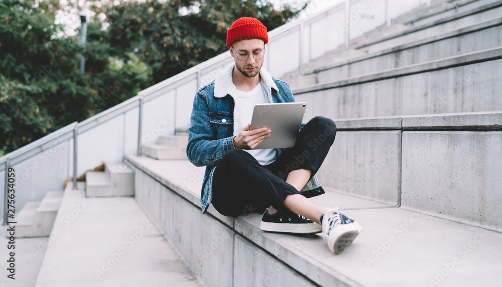 Poster millennial man in casual apparel resting at urban setting searching information on digital tablet, c
