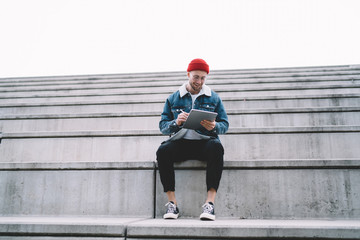 Happy millennial hipster guy typing text message or content publication for social networks while sitting at city stairs and connecting to 4g wireless, concept of digital technology for chatting