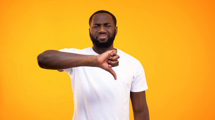 Disappointed afro-american guy showing thumbs down on yellow background, hater