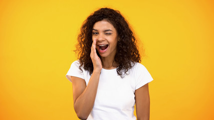 Smiling woman whispering secrets and winking on camera yellow background, rumors