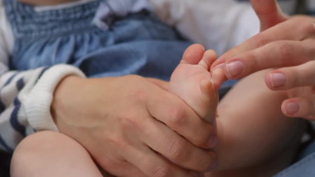 Close-up hands and tiny feet shot of mother counting and gently fondling her little baby daughter’s toes, who is sitting in her mom’s lap
