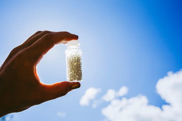 Selective focus on person hand holding glass jar full of small white round homeopathy pills against blue sky background.
