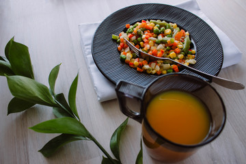 Frozen fried vegetables on black plate. Vegan breakfast with juice. Vegetables food mix on white wooden background with plant.