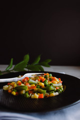 Frozen fried vegetables on black plate. Vegan breakfast. Vegetables food mix on white wooden background with plant.
