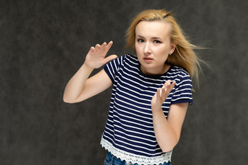 Portrait to the waist of a pretty interesting fashionable blonde girl on a gray background in a striped blue t-shirt. Standing in front of the camera, smiling, showing hands. Shows a lot of emotions.