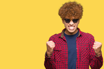 Young handsome man with afro hair wearing sunglasses celebrating surprised and amazed for success with arms raised and open eyes. Winner concept.