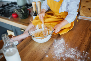 Little girl helping mom bake a cake in the kitchen. Closeup with small cute hands kneading dough in a bowl. Bottle of milk and scattered flour beside.