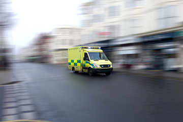 Fototapeta na wymiar Yellow emergency ambulance rushing through a city to the hospital with full flashing blue lights and siren. Speed is emphasized by the motion blurred urban background. Vehicle is generic: no visible m