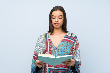 Young woman over isolated blue wall holding and reading a book