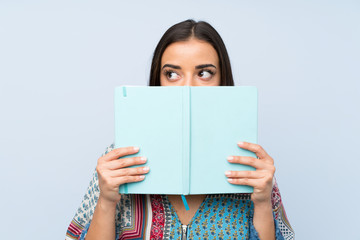 Young woman over isolated blue wall holding and reading a book