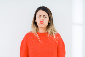 Young woman wearing casual red sweater over isolated background puffing cheeks with funny face. Mouth inflated with air, crazy expression.