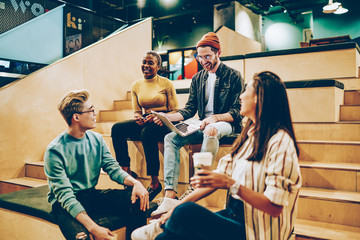 Cheerful young multiracial hipsters laughing at jokes while having meeting for discuss homework project, creative group of male and female colleagues satisfied with friendly atmosphere on job.