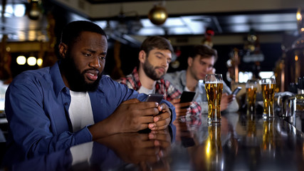 Sad multiethnic men surfing internet on phones in pub instead of communicating