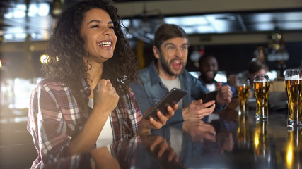 Smiling biracial woman in pub celebrating winning sports bet, bookmaker app