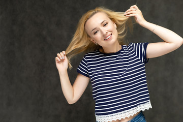 Portrait to the waist of a pretty interesting fashionable blonde girl on a gray background in a striped blue t-shirt. Standing in front of the camera, smiling, showing hands. Shows a lot of emotions.