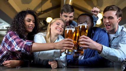 Cheerful friends clinking beer glasses, celebrating team victory in bar, leisure