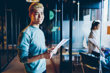 Positive blonde asian male holding paper with good financial report standing in coworking office, smiling man startupper business owner satisfied with occupation and ideas checking  documents