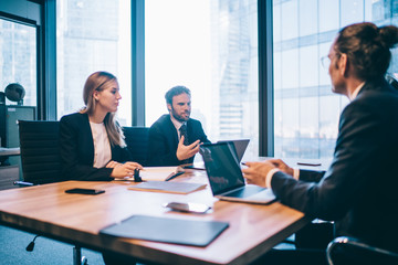 Intelligent professional people cooperating on ideas for financial strategy of company during brainstorming meeting in conference room, serious partners sitting at desktop and consultancy together