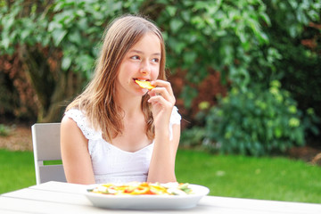 Pretty preteen girl eating dried fruit and vegetable chips in garden outdoors at summer day....