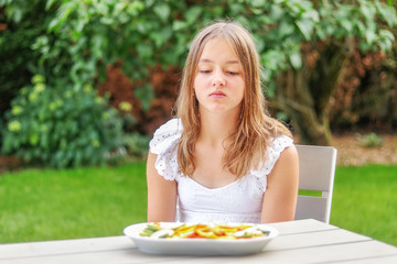 Pretty preteen looking at dried fruit and vegetable chips with unhaddy face sitting in garden outdoors at summer day. Doesn’t like healthy snack. Summer lifestyle