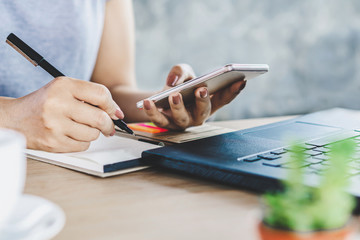 woman hand holding smart phone and pen writing on notebook with laptop on desk