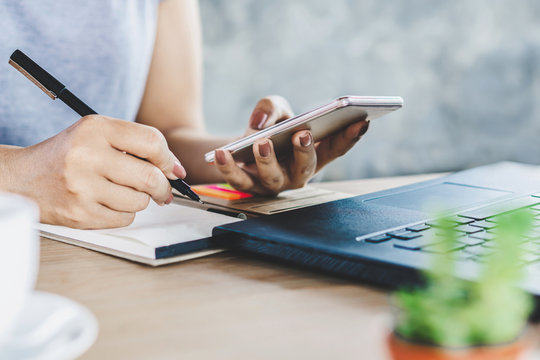 woman hand holding smart phone and pen writing on notebook with laptop on desk