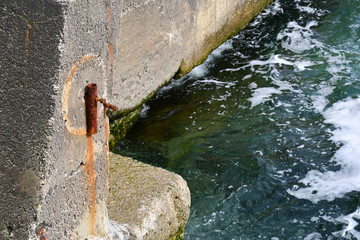 Weathered concrete blocks of pier with old rusty construction near green blue water with wavy texture and white splashes. Black sea shore of Odessa Ukraine. Sea water background. Marine backdrop  