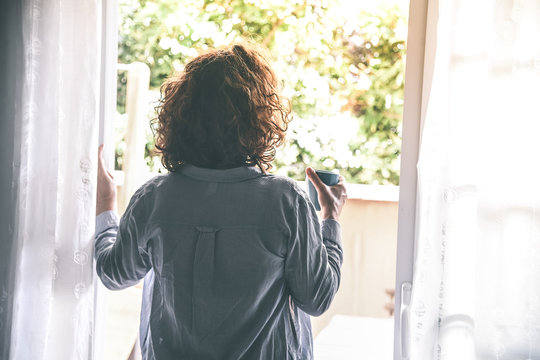 Back view of a young woman drinking a cup of tea looking outside the window at home Curly girl doing breakfast in the soft light of the morning Relax holiday leisure and health and peaceful concept