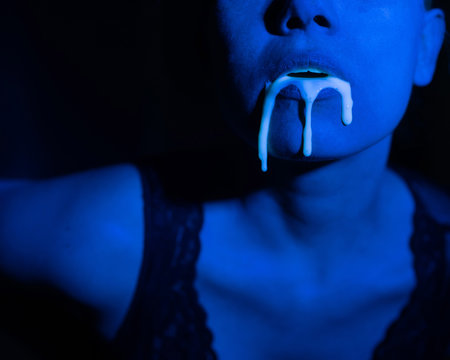 A thick white liquid flows down the lips of a young European girl. Dripping down. Ultraviolet, blue light. Close-up portrait. Spit out.