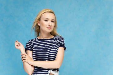 Portrait to the waist of a nice interesting fashionable blonde girl on a blue background in a striped blue t-shirt. Standing in front of the camera, smiling, showing hands. Shows a lot of emotions.