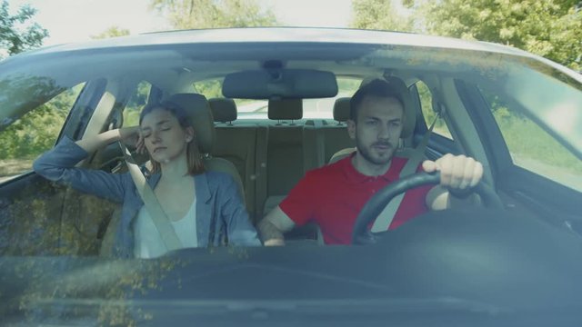 Seriously concentrated young man in red t-shirt driving car. Asleep attractive girl sitting near guy. Daytime. Green nature. Passage. Forest.