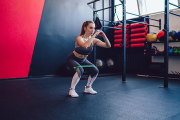 Sporty woman squatting doing sit-ups with resistance band, active Caucasian female in fashionable sportswear training body muscles with elastic equipment, concept of strength and motivation