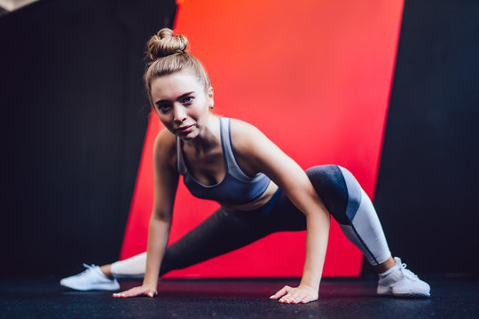 Portrait of beautiful Caucasian bodybuilder in casual tracksuit doing lunge exercise for training legs muscles, sporty hipster girl stretching in pilates studio and looking at camera indoors