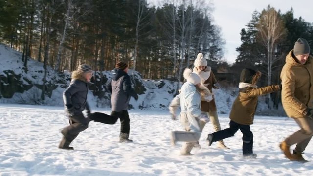 Full shot of happy young family of seven playing action games on cold winter afternoon on snowy frozen over lake, racing, chasing and trying to catch each other, all wearing warm seasonal clothing