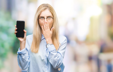 Fototapeta premium Young beautiful blonde business woman showing screen of smartphone over isolated background cover mouth with hand shocked with shame for mistake, expression of fear, scared in silence, secret concept