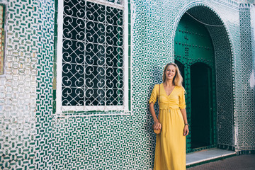 Half length portrait of cheerful woman tourist in yellow sundress posing near bohemian exterior in...
