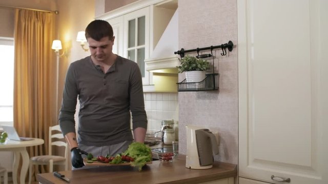 Thigh-up shot of 20-something handicapped man with myoelectric prosthetic forearm carrying cutting board with chopped vegetables over to table to make salad, then picking up mug near kitchen sink