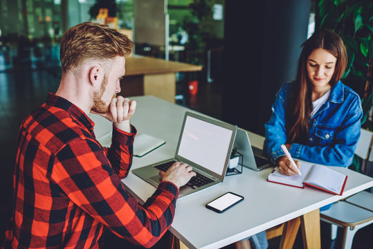Concentrated male it professional working remotely indoors using wireless internet connection for programming on laptop device with blank screen for advertising text sitting at desk with colleague
