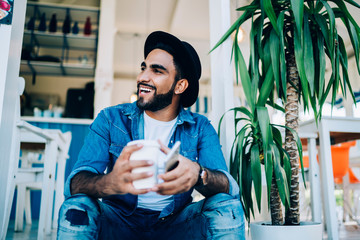 Cheerful Spanish millennial looking away and laughing during leisure time sitting with coffee cup...