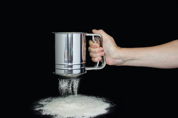 hand holds metal flour sifter, a sieve sprinkled flour. Isolated on black background