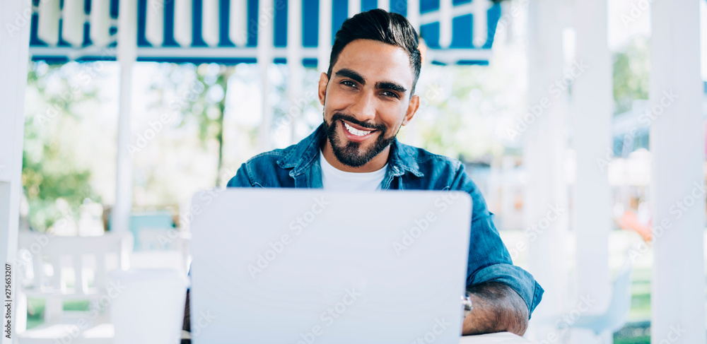Poster portrait of cheerful turkish software developer sitting at desktop with laptop device and smiling at