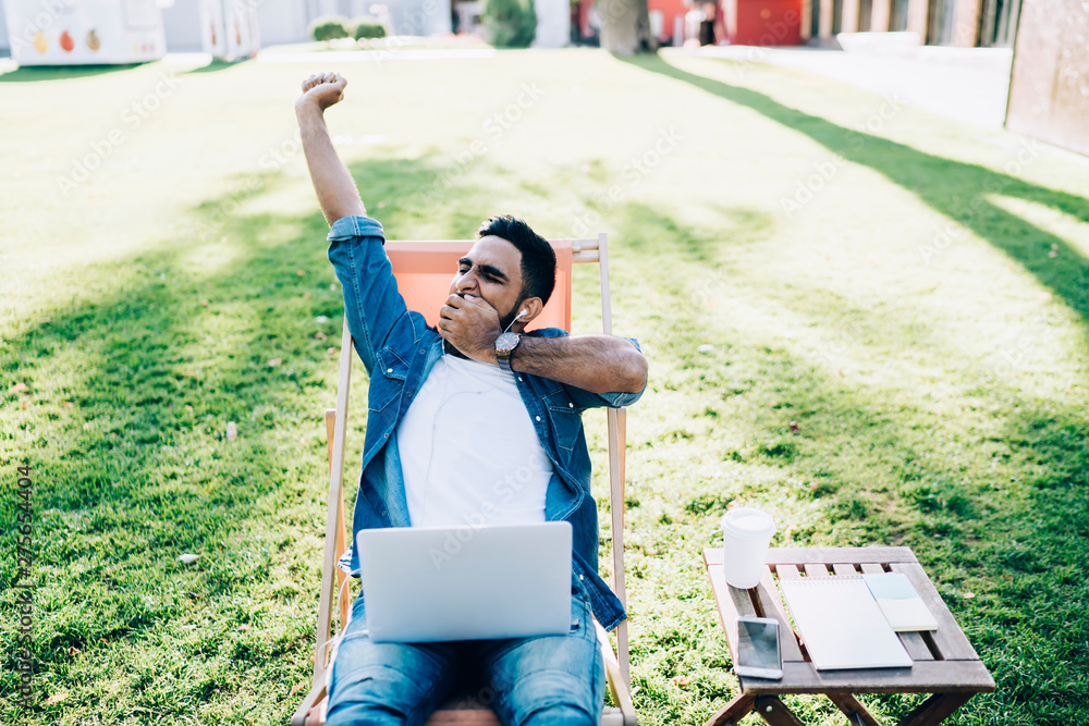 Poster tired male freelancer with closed eyes yawning outdoors while sending time for doing startup project