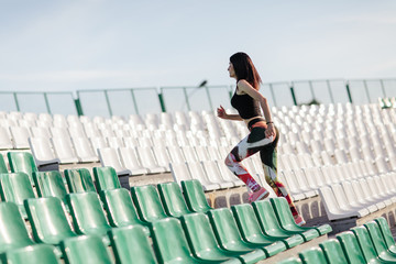 Female athlete with sports figure in leggings and black top running up the stairs of a stadium. Woman in fitness wear doing workout running on stairs beside the seats of a stadium.