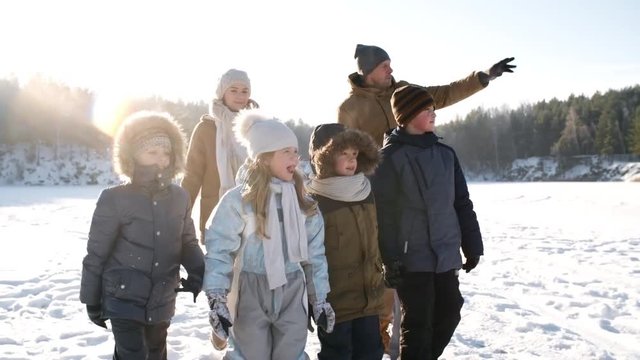 Following medium shot with lens flare of doting young parents and five kids walking in snow across frozen lake on cold winter afternoon in dazzling sunlight, chatting, pointing away and smiling