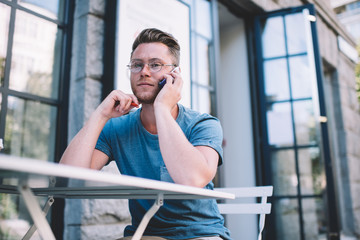 Serious student speaking on cellphone while spending time in street coffee shop, young man in casual wear and eyeglasses phoning for talking concerning news about organisation, communication concept