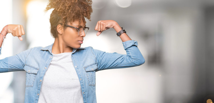 Beautiful young african american woman wearing glasses over isolated background showing arms muscles smiling proud. Fitness concept.