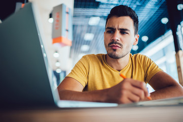Serious male student puzzled on information on website making online analyzing via laptop application, young hipster guy concentrated on university course work spending time for e learning with laptop